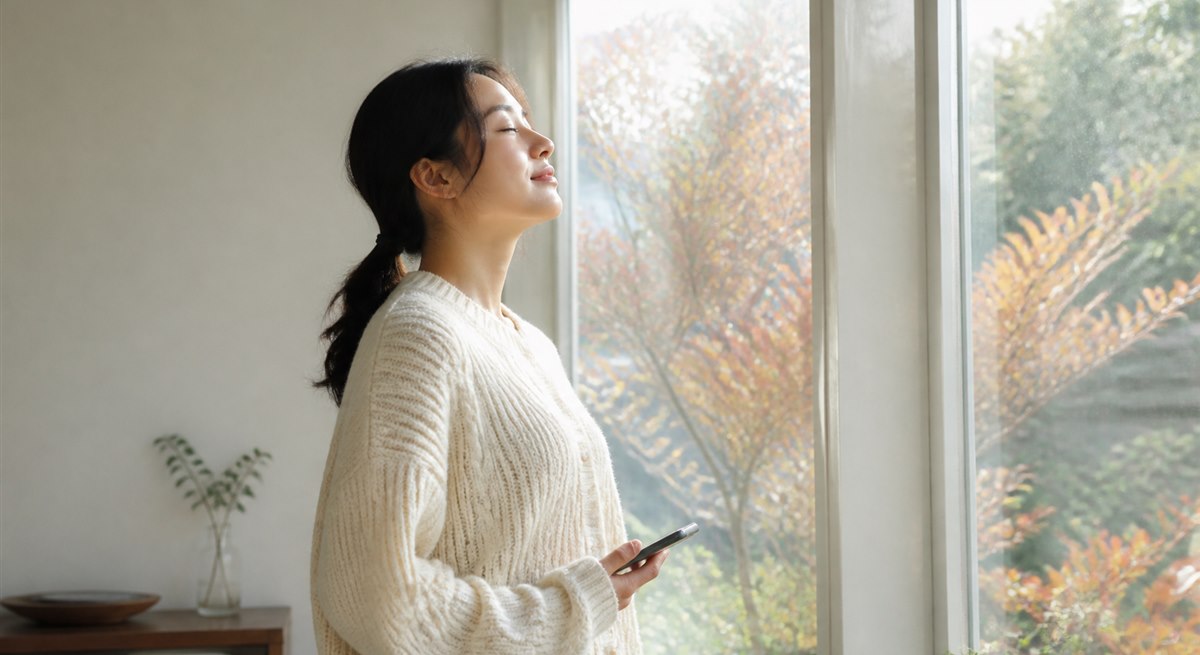 晩秋の窓辺でスマートフォンを手に深呼吸する日本人女性